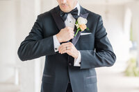 A groom in a black tux adjusts his cuffs while standing with a boutaniere