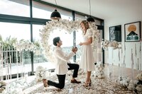 Harry proposing to Karolin indoors surrounded by a white floral arch and rose petals covering the floor, photographed by Auckland proposal photographer Zanthe Vorsatz.