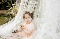 A beautiful toddler with curly hair and striking blue eyes sits amidst sheer white lace fabric outdoors in a garden setting in UK