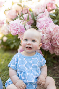 baby girl smiles up at parents (off camera) while sitting in front of bushy pink peony flowers at nichols arb in ann arbor michigan