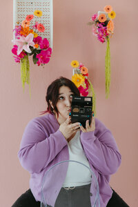 A person with purple hair and red highlights, and a playful expression holds a vintage Polaroid camera up to their lips as if blowing it a kiss. They wear a cozy lilac cardigan over a white top with black pants and gold jewelry. The background features a soft pink wall adorned with colorful hanging floral arrangements in shades of pink, orange, and yellow.