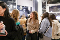 A group of people socializing at an indoor networking event. Two women in the foreground are engaged in conversation, with one gesturing as she talks. Others chat in the background. A table nearby holds snacks and beverages, reflecting a casual and friendly atmosphere typical of Seattle gatherings.