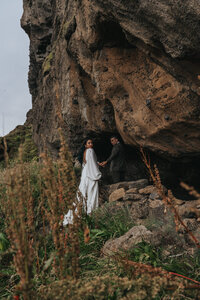 Husband and wife hold hands and peer over their shoulders in a grassy area in Iceland