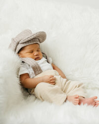 Small baby boy on a white fur backdrop in suspenders and a little old man's cap.