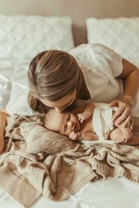 Newborn baby at home, on a bed with mom and she is showering the baby with kisses on her precious cheeks. The are both on a cozy bed with pillows and a soft blanket.