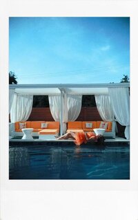 A woman in a coral dress leans back gracefully on a pool’s edge with white cabanas in the background under a clear blue sky.