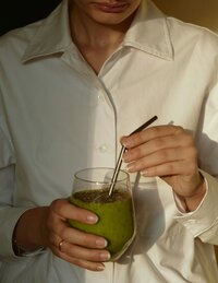 A woman in a white button down shirt holds a glass with a green smoothie in it and a metal straw