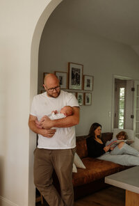 A father stands holding his newborn in an arched doorway of the livingroom as the mother and their two year old read on the couch in the background. They all are wearing timeless neutral colors that exude calm and coziness. These were taken by a fine art New England Photographer. 