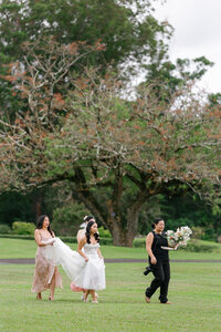 A photographer holding a bouquet of flowers, walking across a field with a newlywed and a wedding guest 
