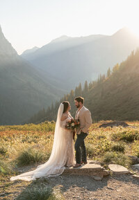 A bride and groom stand together in soft golden light surrounded by Glacier National Park’s mountain peaks and valleys, captured by Sydney Breann Photography during their intimate Montana elopement.