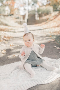 One-year-old baby girl wears a birthday hat as she sits on a knit blanket during an outdoor milestone photo session in Kansas City.