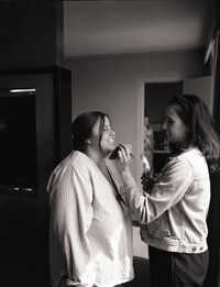 A black-and-white film photograph from a makeup artist's branding session, capturing a candid moment of the artist applying makeup to a smiling client. The relaxed and natural setting showcases the artist's warm and professional approach, emphasizing their ability to create a comfortable and enjoyable experience for clients. The soft lighting and timeless feel reflect the artistry and authenticity of their brand.