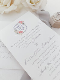 Wedding invitation with blue and coral floral monogram crest and calligraphy.