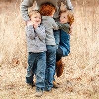 Three young children climb on their dad's legs while he gives them a big hug.