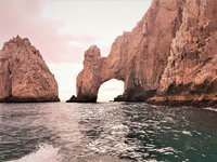 A rugged rock formation with a natural arch rises from the sea at Land's End in Cabo San Lucas, Mexico. Nestled among Mexico all-inclusive resorts, the calm ocean mirrors an overcast sky with a warm, pinkish hue, offering a breathtaking view for travelers seeking serene beauty.