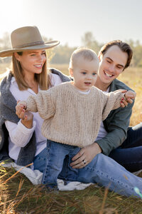 a family sit in a grassy field at sunrise as their baby tries to walk. The baby holds mom's hands and the parents lovingly smile down at the child.