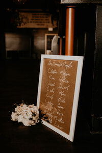 A wedding seating chart written in white ink on brown paper, displayed in a white frame, accompanied by a bouquet of white and blush flowers, resting on a dark wooden floor.