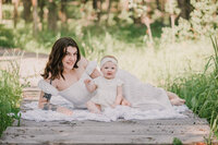 A bright and airy portrait of a mother laying on a wooden boardwalk with her baby daughter sitting in front of her, both smiling.  Captured by Brittany Danielle.