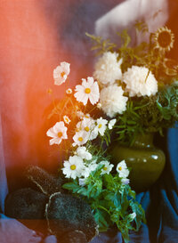 A 35mm film photograph from a florist branding session featuring an artistic floral arrangement with white cosmos, lush greenery, and dried sunflowers. The composition is set against a deep blue fabric backdrop, with a soft orange light leak adding a dreamy, vintage touch. The image highlights the organic beauty and timeless artistry of floral design, perfect for showcasing the creative essence of a florist's brand