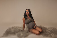 Portrait taken by Atlanta, Georgia maternity photographer of a 35 week pregnant woman dressed in gray sheer maternity dress on gray backdrop posing standing up holding her baby bump and looking to the side