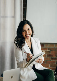 Jessica wearing a white blazer, smiling as she writes in her planner