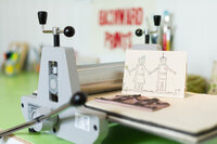 Vignette of a stamped press printing card featuring two robots holding hands and the stamp and press machine for accent branding image.