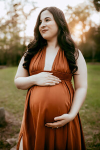 Pregnant woman in a brown dress stands outdoors, cradling her belly with both hands, surrounded by greenery and a soft, glowing sunset.