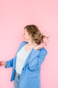 Kat Murphy in a blue blazer flipping her hair in front of a pink background