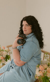 Client of Bostron Design Studio, Brigette is sitting on a chair with pink and white floral arrangements around her. She is facing to the side and holding her camera in one hand.