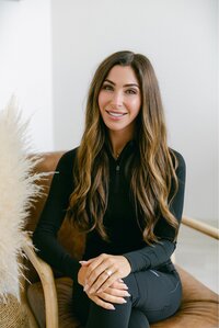 A woman with long brown hair wearing a black outfit sits smiling on a brown chair with her hands clasped, next to a decorative arrangement of light-colored pampas grass at a Maine medical aesthetics clinic.