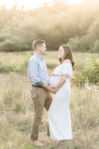 Pregnant woman standing face to face with husband. Both dressed in light neutral colors standing in the middle of a big field with tall grass and wildflowers. Portland Maternity Photographer
