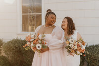 A newlywed couple standing one behind the other smiling as they each hold a bouquet of flowers 