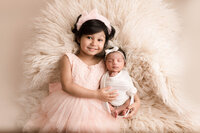 Siblings on cream smiling hugging each other newborn baby and older sibling