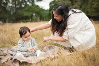 A mother and her one year old daughter sit in the long grass during a late summer family photo shoot in south west London. The mother crouching to stroke the baby's hair.