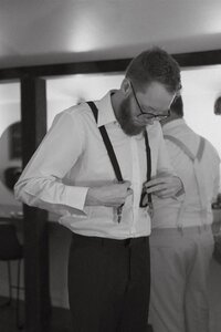 Black and white photo of a groom adjusting his suspenders while getting ready for the wedding at Stone Spring Farms in Pennsylvania — a quiet, candid moment captured before the ceremony.