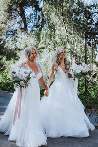 Wedding Photographer, bride and bride hold hands as they walk together, both with their bouquets and happy