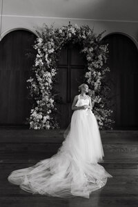 Black and white bridal portrait beneath elegant floral arch, captured in timeless editorial wedding style.