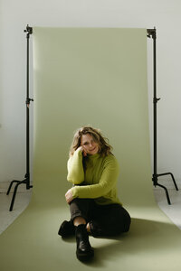 Sara Dobbins sits on the floor in leaning  on her elbow on her knee . She sits in front of a green photo backdrop