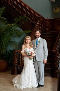 Bride in white lace mermaid wedding dress and groom in light gray suit pose together at the White Room in historic downtown Saint Augustine, standing in front of an ornate wooden staircase with decorative carved railings and large tropical palm plants