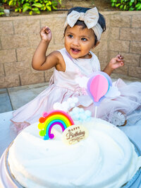 A one-year-old birthday girl in a pale drink dress and bow smiles with her arms in the air behind her white birthday cake, which is decorated with a rainbow, clouds, and hot-air balloon. Photo by SAVI Photography - Los Angeles CA Photographer
