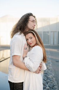 A man holds his fiance against his chest while she looks at the camera with a soft smile. They're in downtown Raleigh.