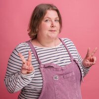 Person called Lucy has shoulder-length brown mousey hair in a navy-and-white striped top and mauve dungarees stands against a pink backdrop, holding up double peace signs with a playful, slightly skeptical expression.
