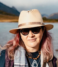 Sonia, a pink-haired, 50-something white woman in a broad-brimmed hat and sunglasses, mountains and a lake in the background