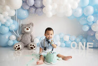 Little boy sitting on a white floor eating cake with blue, grey and white balloons for his Burlington, Ontario cake smash photography session.
