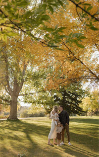 A husband and wife kiss while holding their baby. Their toddler stands at their feet. They have trees all around them.