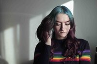 A young, feminine presenting person with blue and purple hair, looks down toward the ground, running their right hand through their hair, as if lost in thought. They are not smiling, and look sad, despite not facing the camera. Their black sweatshirt has horizontal, rainbow stripes across the top.