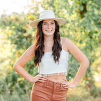 A high school senior wearing a white hat and fringe top puts her hands on her hips and smiles for the camera.