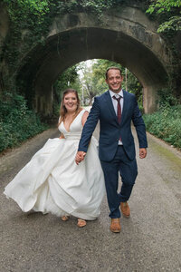 Bride and groom walk holding hands and laughing in front of stone tunnel
