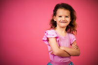 a child wearing a pink top with sassy smile crosses her arms and smiles for this children's portrait on bright pink.
