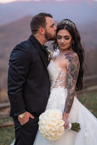 punk bride and groom stand on a mountain top at sunset. groom in full black tux gently kisses bride on forehead. bride wears a diamond crown hair piece and lace open back wedding gown while showcasing her beautiful black hair and full color tattoo sleeve on her arm